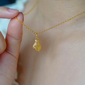 Pode incluir: Colar dourado com um pendente texturizado em forma de gota. O pendente pende de uma corrente delicada. O colar é exibido sobre um fundo neutro, realçando a cor dourada.