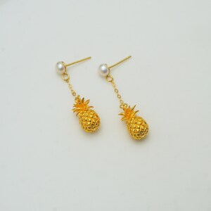 May include: A pair of gold-toned drop earrings featuring a small white pearl stud at the top, connected by a thin gold chain to a textured, three-dimensional pineapple charm. The pineapple charms are detailed with a cross-hatched surface pattern.