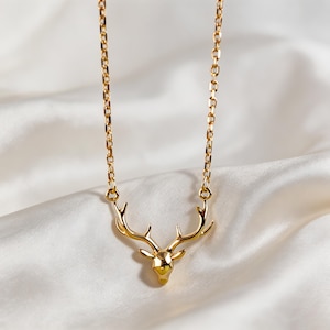 Puede incluir: Un collar de tono dorado que presenta un colgante de cabeza de ciervo pulida con astas prominentes. El colgante está suspendido de una delicada cadena de tono dorado con eslabones ovalados, mostrando un acabado metálico liso y brillante.