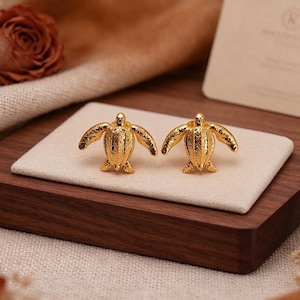 May include: A pair of gold-toned stud earrings shaped like sea turtles. Each earring features a textured shell and flipper design with a polished metallic finish, showcasing a detailed, symmetrical form for both the left and right sides.