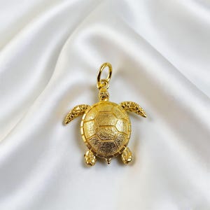 May include: A gold-coloured turtle pendant with a textured shell and detailed flippers and head. The pendant has a loop at the top for a chain. The turtle is displayed on a white, silky background.