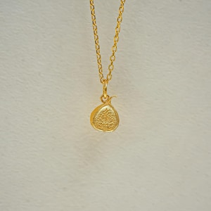 May include: A gold-toned necklace featuring a delicate chain and a small, textured fig-shaped pendant. The pendant displays a detailed, seed-like pattern on its flat surface, suspended from a simple jump ring attached to the thin metal chain.