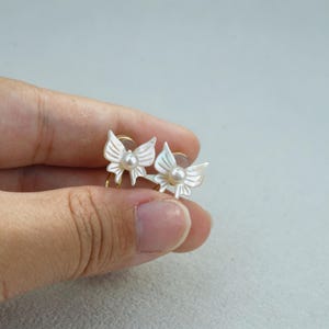 Puede incluir: Un par de pendientes con forma de mariposa que presentan alas de nácar blanco iridiscente y una pequeña perla central. Los pendientes están fabricados con herrajes de metal dorado e incluyen un mecanismo de cierre circular para su sujeción.