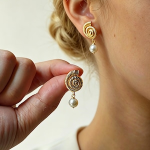 Op de afbeelding: Goudkleurige oorbellen met een spiraalvormig ontwerp en een bungelende witte parel. Eén oorbel wordt gedragen, de andere wordt in de hand gehouden. De oorbellen hebben een getextureerd, geribbeld oppervlak.