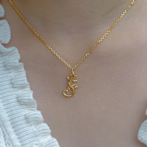 Puede incluir: Un collar de tono dorado que presenta una cadena delicada y un pequeño colgante detallado con forma de lagarto. El colgante está suspendido del centro de la cadena, mostrando un acabado liso y pulido que refleja la luz con brillo.