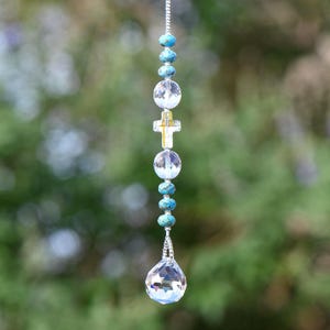 May include: A hanging crystal suncatcher with a silver chain. The suncatcher features clear, round crystal beads, turquoise beads, and a gold-colored cross. A large, faceted crystal ball hangs at the bottom.