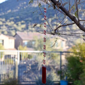 Puede incluir: Un carill&oacute;n de vidrio rojo colgando de una rama de &aacute;rbol. El carill&oacute;n tiene una larga cadena de cuentas rojas y un tubo de vidrio rojo en la parte inferior.