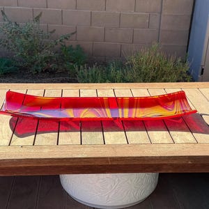 Puede incluir: Una bandeja rectangular larga de vidrio con un patrón de remolinos vibrantes en colores rojo brillante, naranja y amarillo. La bandeja tiene bordes ligeramente curvados hacia arriba y un acabado liso y brillante que refleja la luz.