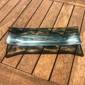 May include: Rectangular glass serving dish with a teal and silver design. The dish has slightly curved edges and a textured appearance, reflecting light. The glass appears to be transparent, allowing the wooden surface beneath to be partially visible.