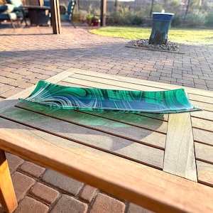 Puede incluir: Una bandeja rectangular de vidrio con un vibrante patrón jaspeado en verde y blanco que imita la apariencia de la piedra de malaquita. La bandeja tiene una forma alargada ligeramente curva con bordes lisos y un acabado de superficie brillante.