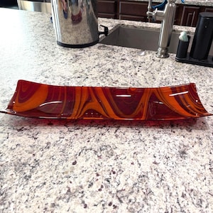 Puede incluir: Una bandeja rectangular larga de vidrio con un patrón de remolinos jaspeados en tonos rojo intenso, naranja quemado y marrón oscuro. La bandeja tiene bordes ligeramente elevados y un acabado suave y brillante que refleja la luz en su superficie.