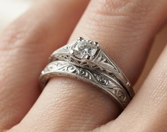 May include: A silver-toned engagement ring set featuring a central round-cut stone and a matching band. Both rings display intricate engraved scrollwork patterns along the metal shanks, showcasing a detailed filigree design throughout the bands.