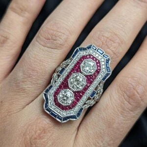 Può includere: Un anello di forma rettangolare con tre diamanti centrali a taglio rotondo incastonati in un bordo di pietre preziose rosse. Il pezzo è incorniciato da un bordo di piccoli zaffiri blu a taglio baguette e numerosi minuscoli diamanti bianchi.