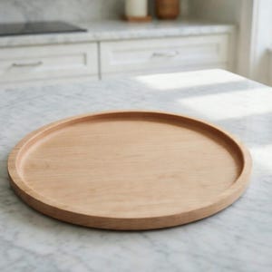 May include: A round, light-colored wooden tray with a raised edge. The tray is made of natural wood and sits on a marble countertop. The tray is approximately 12 inches in diameter and is suitable for serving food or displaying items.