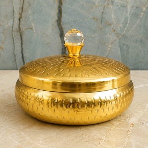 Peut inclure: Un récipient rond en métal doré avec un couvercle assorti. La surface du récipient et du couvercle présente un motif gaufré répétitif. Un bouton en cristal transparent, facetté et en forme de diamant, surmonte le centre du couvercle.
