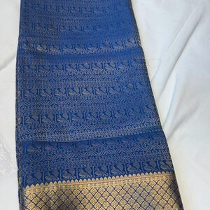 May include: A vibrant blue fabric featuring intricate gold-toned woven patterns. The design includes horizontal bands of repeating peacock motifs and geometric borders, with a wide, ornate gold-toned decorative hem at the bottom edge.