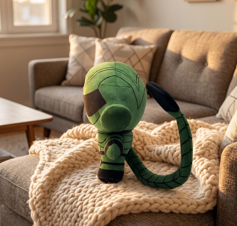 Op de afbeelding: Een groen pluche speelgoed met een zwart vizier en een gebogen staart. Het speelgoed staat op een cr&egrave;mekleurige gebreide deken, met een bank op de achtergrond. Het speelgoed heeft een uniek ontwerp en is gemaakt van zacht materiaal.