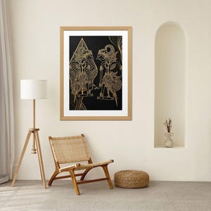Puede incluir: Una obra de arte enmarcada con dos figuras doradas sobre un fondo negro. Las figuras tienen tocados y trajes detallados. La obra de arte se exhibe en una habitación con una silla de madera, una lámpara de pie y un pequeño otomán tejido.