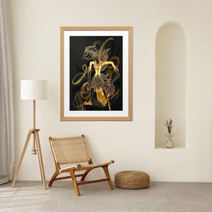 Op de afbeelding: Ingelijste kunstdruk met een gouden illustratie van een traditionele Indonesische Wayang Kulit-pop op een zwarte achtergrond. Het kunstwerk zit in een houten lijst en wordt getoond in een kamer met een stoel en een lamp.