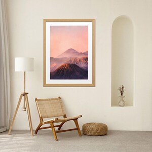 Puede incluir: Una impresión enmarcada de un paisaje montañoso bajo un cielo rosa y púrpura. La impresión está en un marco de madera clara. Una silla de madera con asiento y respaldo tejido, una lámpara de pie y un pequeño otomán tejido están en primer plano.