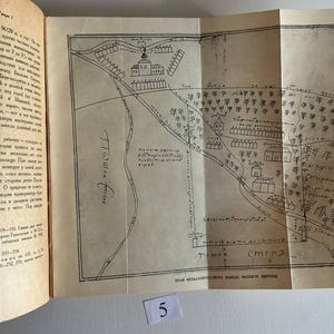 K&ouml;nnte beinhalten: Ein aufgeschlagenes Buch mit cremefarbenen Seiten, das auf der rechten Seite eine schwarz-wei&szlig;e Strichzeichnung einer historischen Karte zeigt. Die Karte stellt Geb&auml;ude, B&auml;ume und Gel&auml;nde mit handschriftlichem kyrillischem Text dar. Ein kleines wei&szlig;es Papieretikett mit der Zahl 5 ist sichtbar.