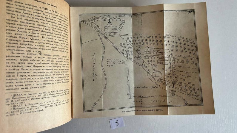 K&ouml;nnte beinhalten: Ein aufgeschlagenes Buch mit cremefarbenen Seiten, das auf der rechten Seite eine schwarz-wei&szlig;e Strichzeichnung einer historischen Karte zeigt. Die Karte stellt Geb&auml;ude, B&auml;ume und Gel&auml;nde mit handschriftlichem kyrillischem Text dar. Ein kleines wei&szlig;es Papieretikett mit der Zahl 5 ist sichtbar.