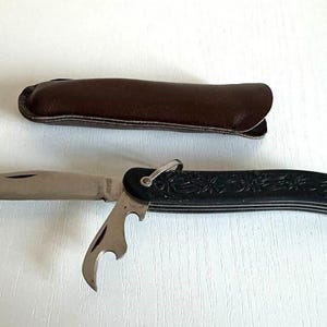 Può includere: Un coltello tascabile con una lama in acciaio inossidabile e un manico nero con dettagli floreali. È dotato di un apribottiglie e di un piccolo anello. È inclusa anche una custodia in pelle marrone.