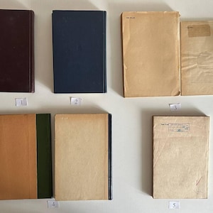 K&ouml;nnte beinhalten: Eine Sammlung von sieben rechteckigen B&uuml;chern, die in zwei Reihen angeordnet sind. Die B&uuml;cher haben verschiedene einfarbige Einb&auml;nde in Dunkelbraun, Marineblau und Beige. Einige Einb&auml;nde weisen Gebrauchsspuren, Papieretiketten und Stempel auf.