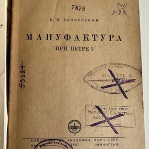 K&ouml;nnte beinhalten: Ein beigefarbener, alter Bucheinband mit schwarzem kyrillischem Text, der "АКАДЕМИЯ НАУК СОЮЗА ССР", "Е. И. ЗАОЗЕРСКАЯ" und "МАНУФАКТУРА ПРИ ПЕТРЕ I" lautet. Der Einband weist mehrere violette Bibliotheksstempel, handschriftliche Zahlen und einen roten Haken auf.