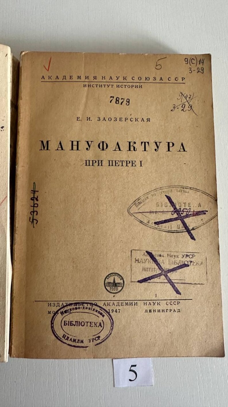K&ouml;nnte beinhalten: Ein beigefarbener, alter Bucheinband mit schwarzem kyrillischem Text, der "АКАДЕМИЯ НАУК СОЮЗА ССР", "Е. И. ЗАОЗЕРСКАЯ" und "МАНУФАКТУРА ПРИ ПЕТРЕ I" lautet. Der Einband weist mehrere violette Bibliotheksstempel, handschriftliche Zahlen und einen roten Haken auf.