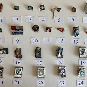 May include: A collection of vintage pins and badges, each with unique designs and colors. The pins are arranged on a white surface, with numbered labels below each one. The collection includes pins with flags, portraits, and other symbols.