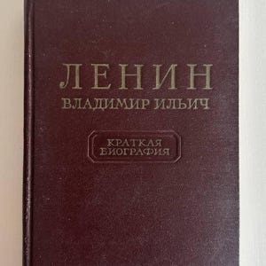 K&ouml;nnte beinhalten: Ein dunkelweinrotes Hardcover-Buch mit goldener Schrift auf dem Einband. Der Text lautet "ЛЕНИН", "ВЛАДИМИР ИЛЬИЧ" und "КРАТКАЯ БИОГРАФИЯ" innerhalb eines rechteckigen Rahmens. Ein kleines wei&szlig;es Papieretikett mit der Zahl "1" befindet sich unten mittig.