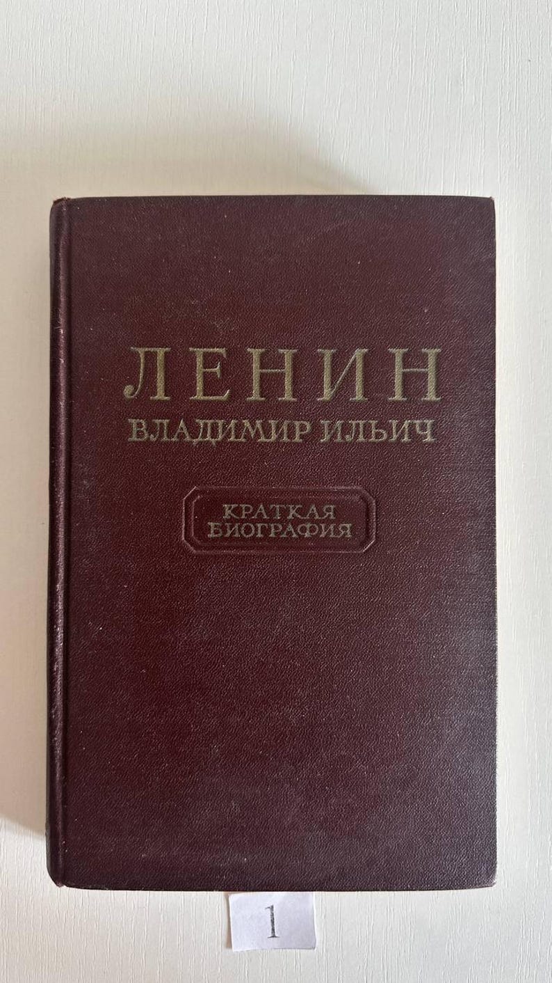 K&ouml;nnte beinhalten: Ein dunkelweinrotes Hardcover-Buch mit goldener Schrift auf dem Einband. Der Text lautet "ЛЕНИН", "ВЛАДИМИР ИЛЬИЧ" und "КРАТКАЯ БИОГРАФИЯ" innerhalb eines rechteckigen Rahmens. Ein kleines wei&szlig;es Papieretikett mit der Zahl "1" befindet sich unten mittig.