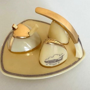 May include: A vintage cream-colored tea set with gold accents. The set includes a sugar bowl, creamer, and a salt shaker with a black and white illustration of a building and the word "Riga". The set sits on a matching tray.