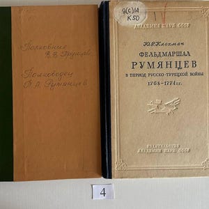 K&ouml;nnte beinhalten: Zwei gebundene B&uuml;cher nebeneinander. Das linke Buch hat einen beigen Einband mit gr&uuml;nem Buchr&uuml;cken und handschriftlichem Text. Das rechte Buch hat einen beigen Einband mit dunkelblauem Buchr&uuml;cken, kyrillischem Text und einem Aufkleber.
