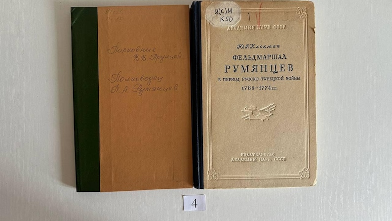 K&ouml;nnte beinhalten: Zwei gebundene B&uuml;cher nebeneinander. Das linke Buch hat einen beigen Einband mit gr&uuml;nem Buchr&uuml;cken und handschriftlichem Text. Das rechte Buch hat einen beigen Einband mit dunkelblauem Buchr&uuml;cken, kyrillischem Text und einem Aufkleber.