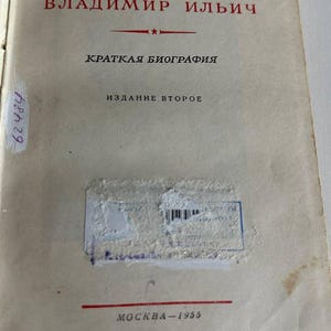 K&ouml;nnte beinhalten: Eine Vintage-Buchtitelseite mit dem Text LЕНИН ВЛАДИМИР ИЛЬИЧ, КРАТКАЯ БИОГРАФИЯ, ИЗДАНИЕ ВТОРОЕ und MOCKBA-1955. Die Seite enth&auml;lt rote und schwarze Tinte, einen rosa Klebestreifen, handschriftliche Zahlen und einen blauen Stempel mit Barcode.