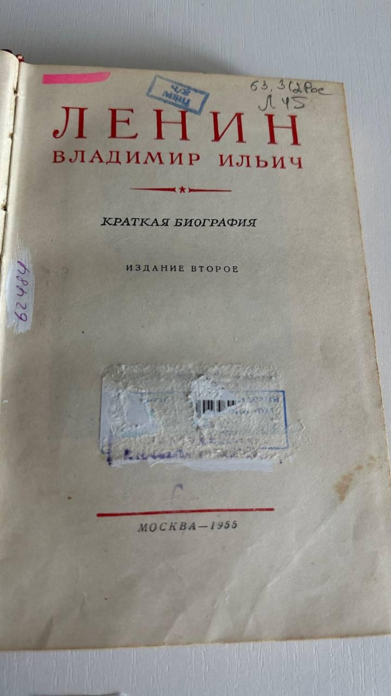 K&ouml;nnte beinhalten: Eine Vintage-Buchtitelseite mit dem Text LЕНИН ВЛАДИМИР ИЛЬИЧ, КРАТКАЯ БИОГРАФИЯ, ИЗДАНИЕ ВТОРОЕ und MOCKBA-1955. Die Seite enth&auml;lt rote und schwarze Tinte, einen rosa Klebestreifen, handschriftliche Zahlen und einen blauen Stempel mit Barcode.
