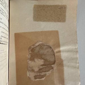 K&ouml;nnte beinhalten: Ein aufgeschlagenes Buch mit cremefarbenen Seiten und kyrillischem Text auf der linken Seite. Auf der rechten Seite befindet sich ein rechteckiges St&uuml;ck transparentes Papier, das einen dunklen, unregelm&auml;&szlig;igen, braunen Fleck bedeckt.