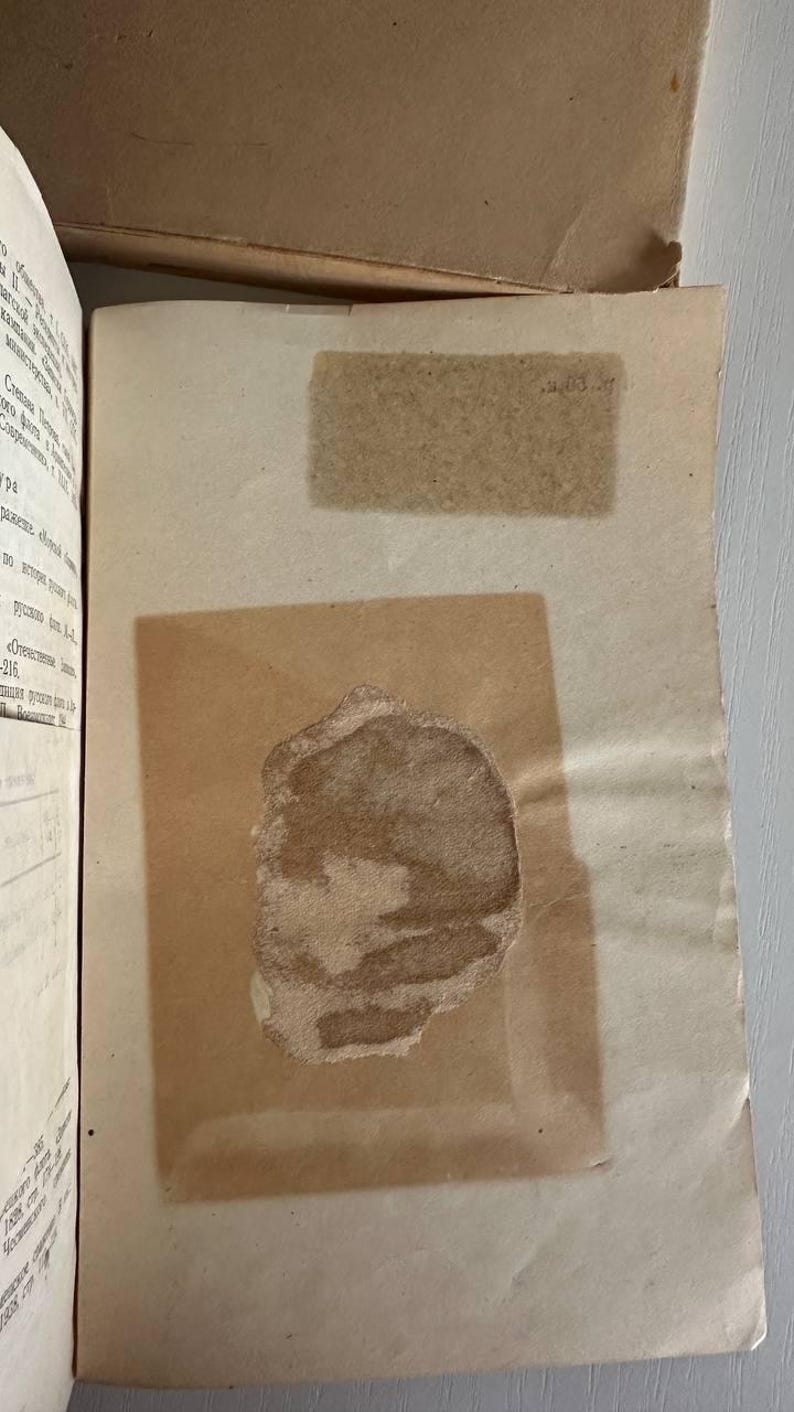 K&ouml;nnte beinhalten: Ein aufgeschlagenes Buch mit cremefarbenen Seiten und kyrillischem Text auf der linken Seite. Auf der rechten Seite befindet sich ein rechteckiges St&uuml;ck transparentes Papier, das einen dunklen, unregelm&auml;&szlig;igen, braunen Fleck bedeckt.