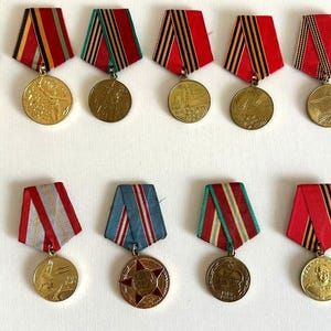 May include: A selection of vintage medals with red, black, white, and blue ribbons. The medals are gold and silver-toned, with diverse designs and inscriptions. The ribbons are striped and solid, some with a star design.