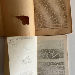 K&ouml;nnte beinhalten: Zwei aufgeschlagene Seiten eines alten russischsprachigen Buches mit beigem Papier. Die obere Seite weist einen gro&szlig;en Riss am linken Rand und gedruckten Text auf. Die untere Seite zeigt gedruckten Text, handschriftliche Notizen und Stempel.