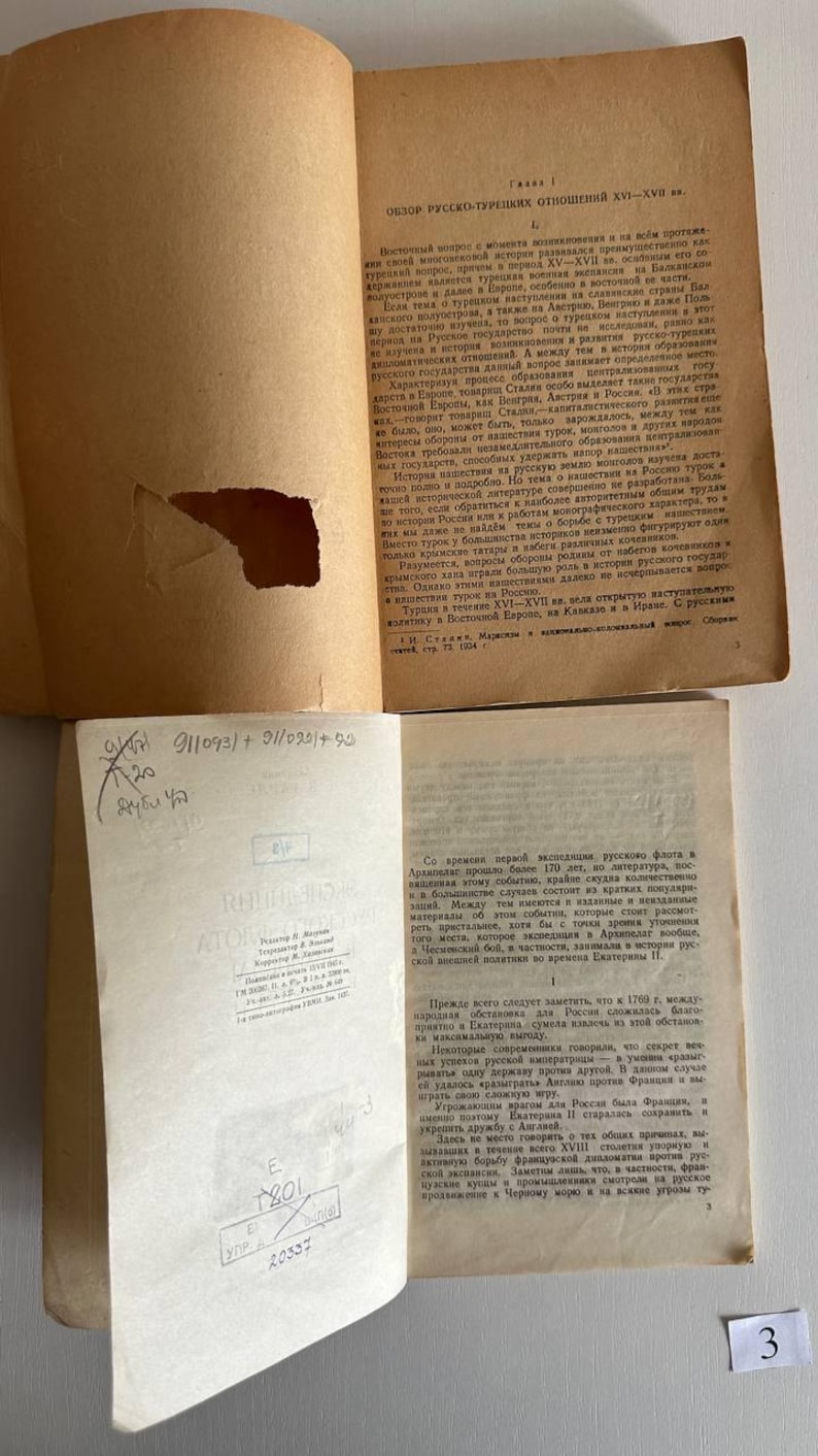 K&ouml;nnte beinhalten: Zwei aufgeschlagene Seiten eines alten russischsprachigen Buches mit beigem Papier. Die obere Seite weist einen gro&szlig;en Riss am linken Rand und gedruckten Text auf. Die untere Seite zeigt gedruckten Text, handschriftliche Notizen und Stempel.
