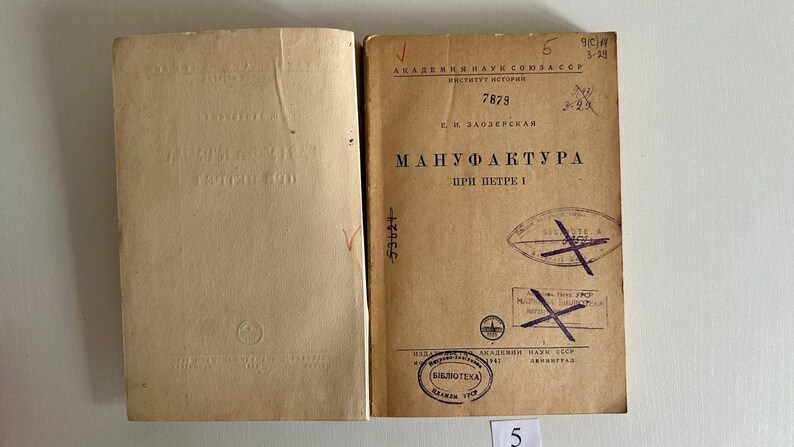K&ouml;nnte beinhalten: Ein aufgeschlagenes antikes Buch mit beigen Seiten. Die rechte Seite zeigt kyrillischen Text, darunter den Titel "МАНУФАКТУРА ПРИ ПЕТРЕ I", handschriftliche Zahlen und mehrere violette Stempel mit durchgestrichenen Markierungen. Die linke Seite ist leer mit einem Logo.