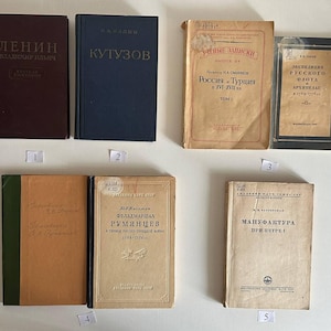 K&ouml;nnte beinhalten: Eine Sammlung von sechs alten B&uuml;chern mit verschiedenen Einb&auml;nden, darunter ein dunkelbraunes Buch mit dem Titel Lenin, ein blaues Buch mit dem Titel Kutusow sowie mehrere beige und schwarze historische B&auml;nde mit russischem Text, angeordnet in zwei Reihen mit kleinen nummerierten Etiketten.