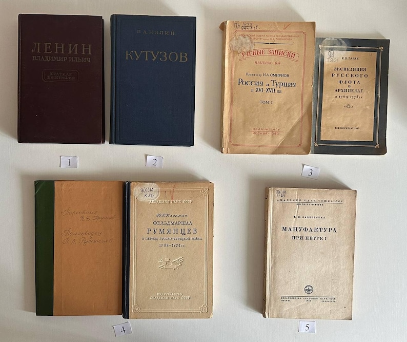K&ouml;nnte beinhalten: Eine Sammlung von sechs alten B&uuml;chern mit verschiedenen Einb&auml;nden, darunter ein dunkelbraunes Buch mit dem Titel Lenin, ein blaues Buch mit dem Titel Kutusow sowie mehrere beige und schwarze historische B&auml;nde mit russischem Text, angeordnet in zwei Reihen mit kleinen nummerierten Etiketten.
