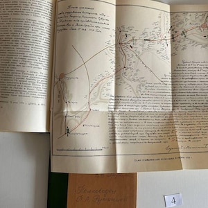 K&ouml;nnte beinhalten: Ein aufgeschlagenes Hardcover-Buch mit cremefarbenen Seiten und dichtem schwarzem kyrillischem Text. Eine gro&szlig;e, gefaltete, hellbraune Karteneinlage ist an der Seite befestigt und zeigt detaillierte topografische Linien, handschriftliche Notizen und eine Skala.