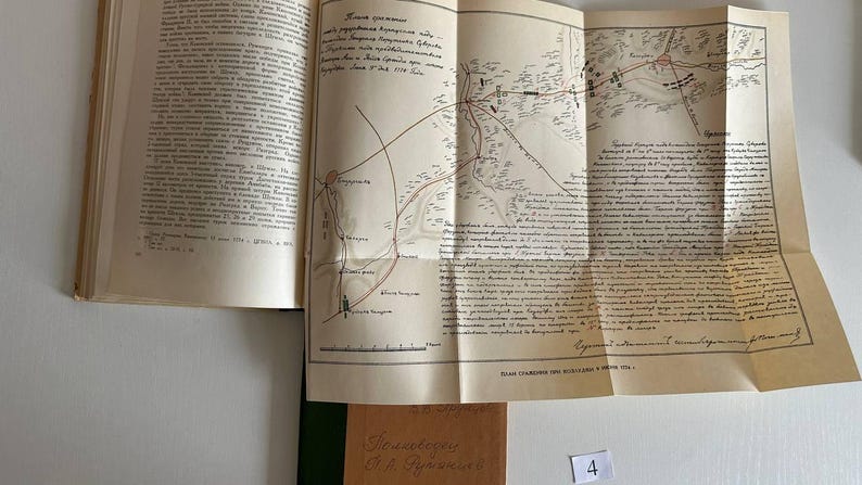 K&ouml;nnte beinhalten: Ein aufgeschlagenes Hardcover-Buch mit cremefarbenen Seiten und dichtem schwarzem kyrillischem Text. Eine gro&szlig;e, gefaltete, hellbraune Karteneinlage ist an der Seite befestigt und zeigt detaillierte topografische Linien, handschriftliche Notizen und eine Skala.