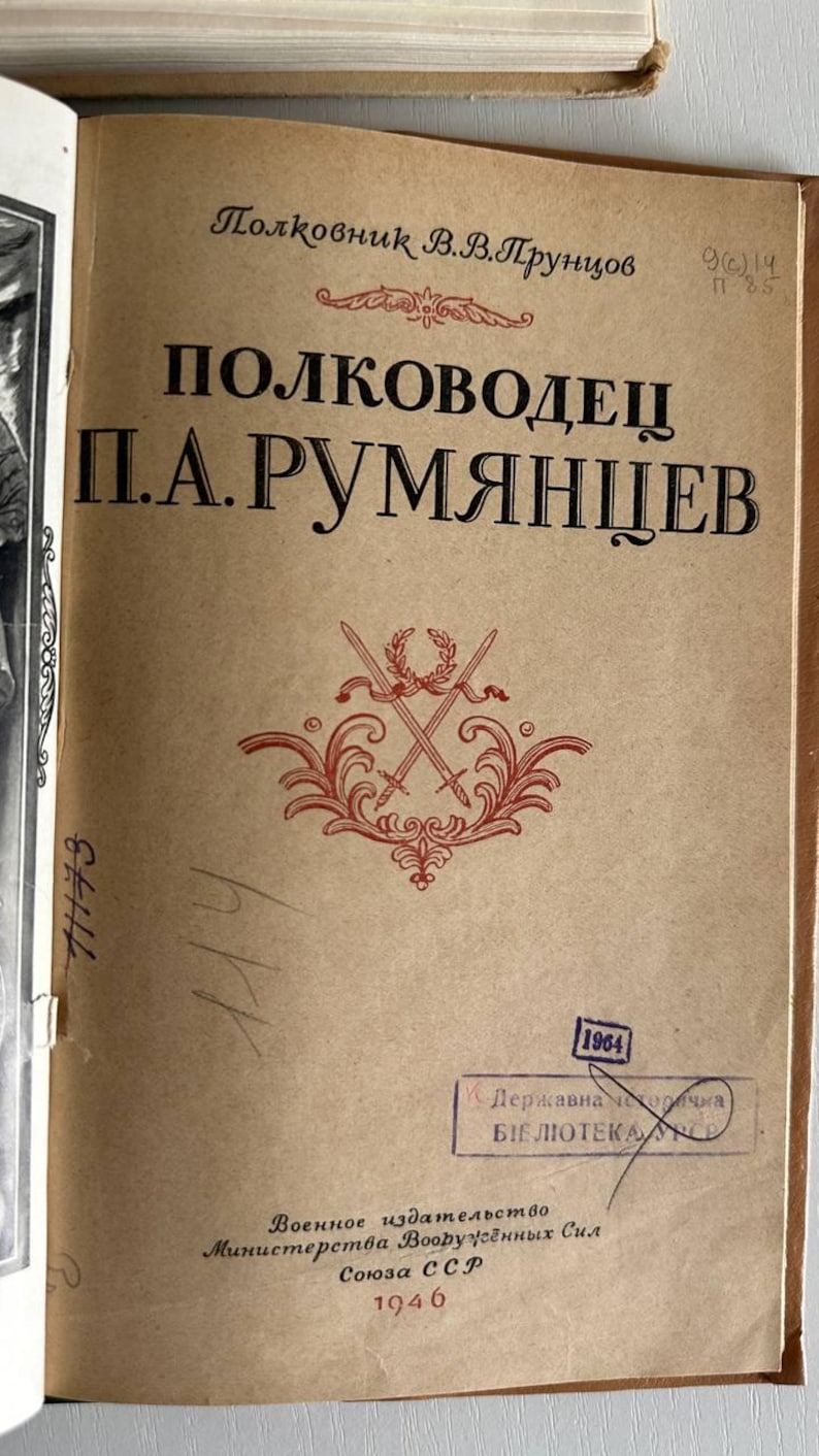 K&ouml;nnte beinhalten: Eine Buch-Titelseite auf beigem Papier mit dem russischen Text "Полковник В.В.Прунцов", "ПОЛКОВОДЕЦ П.А.РУМЯНЦЕВ" und "1946". Ein rotes dekoratives Emblem mit gekreuzten Schwertern und einem Lorbeerkranz befindet sich &uuml;ber einem blauen Bibliotheksstempel.