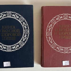 Könnte beinhalten: Zwei Hardcover-Bücher mit dem Titel "Книга о вкусной и здоровой пище" in goldener Schrift. Ein Buch ist marineblau, das andere weinrot. Beide Bücher haben ein weißes kreisförmiges Design auf dem Einband. Das marineblaue Buch hat die Nummer 1.